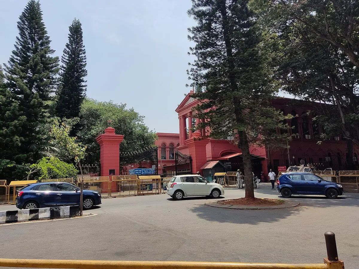 Le bâtiment rouge de la Haute Cour du Karnataka derrière de grands arbres, des voitures sur la route