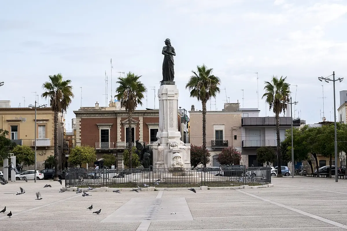 Une place de quartier à Bari avec un monument, des palmiers et des façades résidentielles