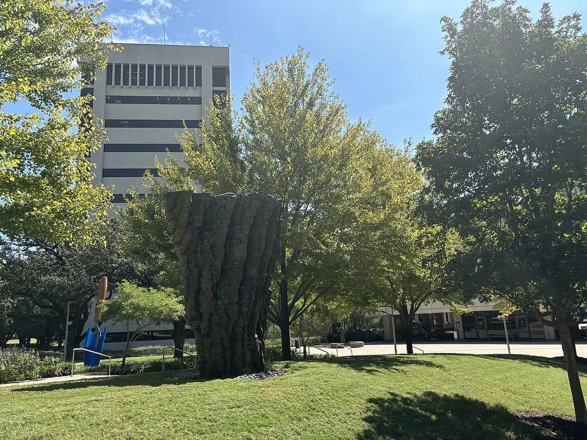 A small park in Dallas with a mature tree strung with lights and a dark abstract sculpture