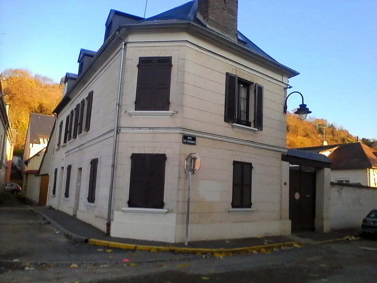 Maison blanche à volets bruns à un angle de rue à Évreux, arbres d’automne derrière