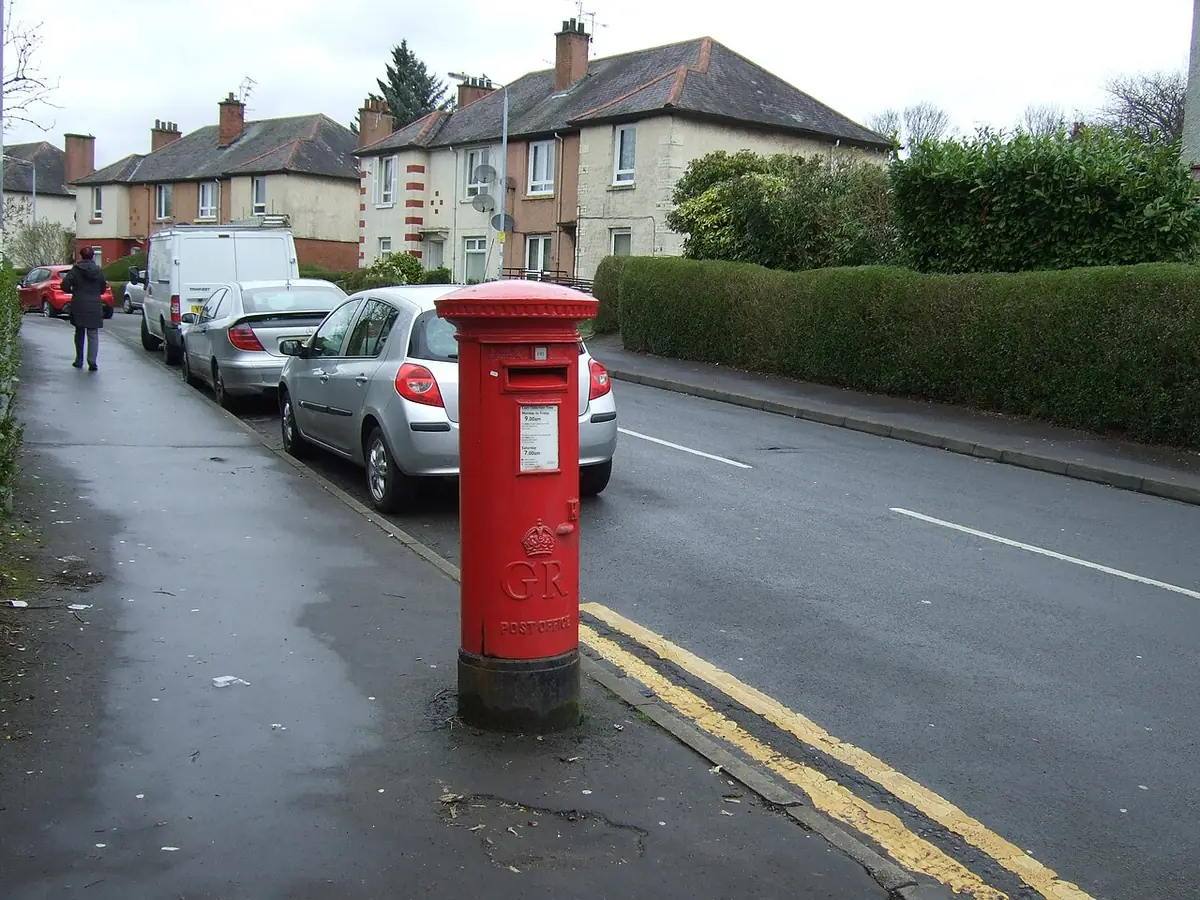 Une rue résidentielle avec des haies, une boîte aux lettres rouge et des voitures garées
