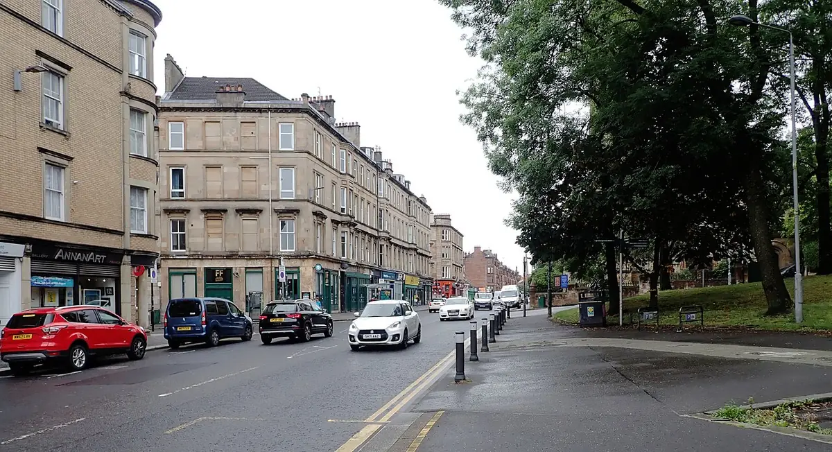 Une rue de Glasgow bordée d’arbres avec des immeubles en grès et des commerces en bas
