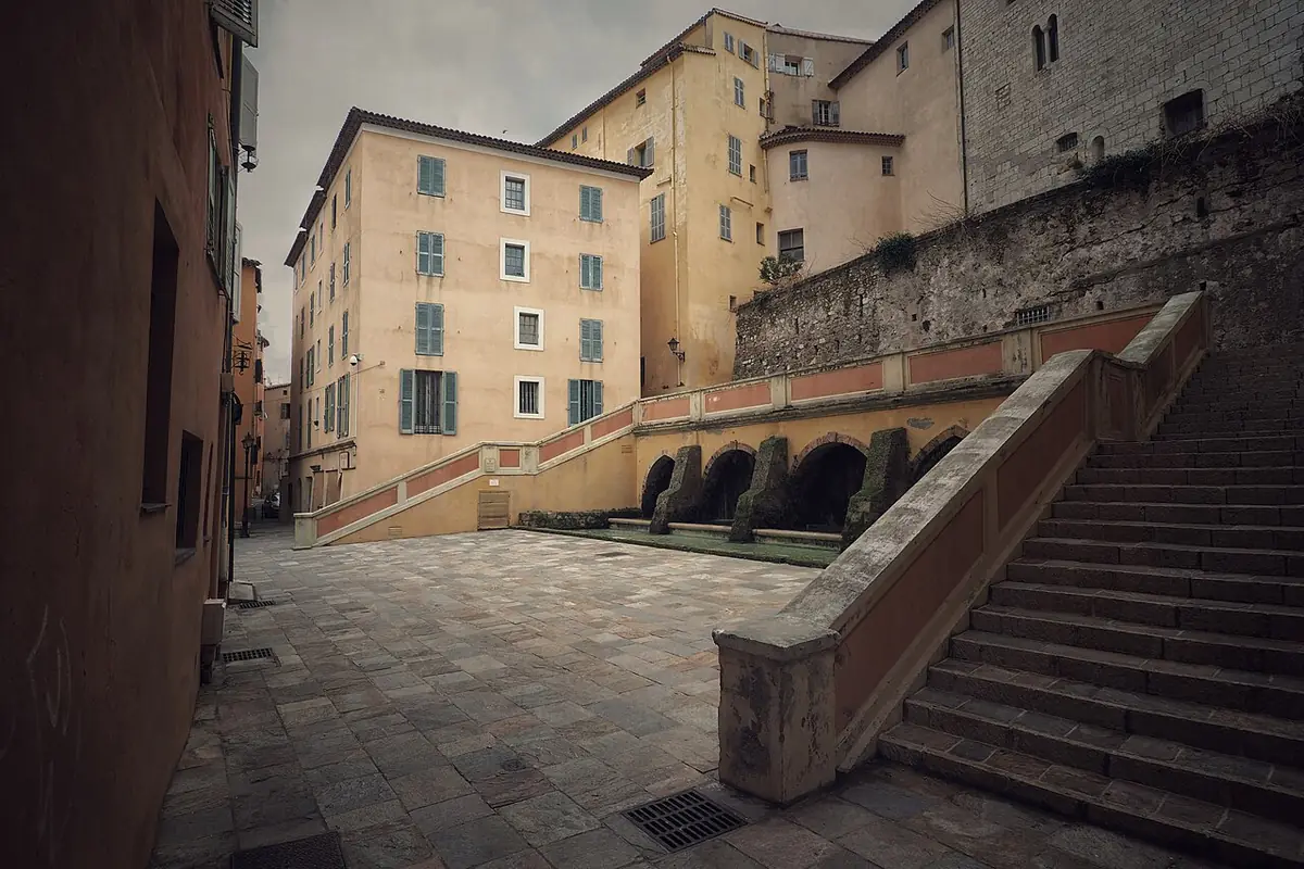 Place pavée et escalier de pierre entre de hautes façades ocre à Grasse