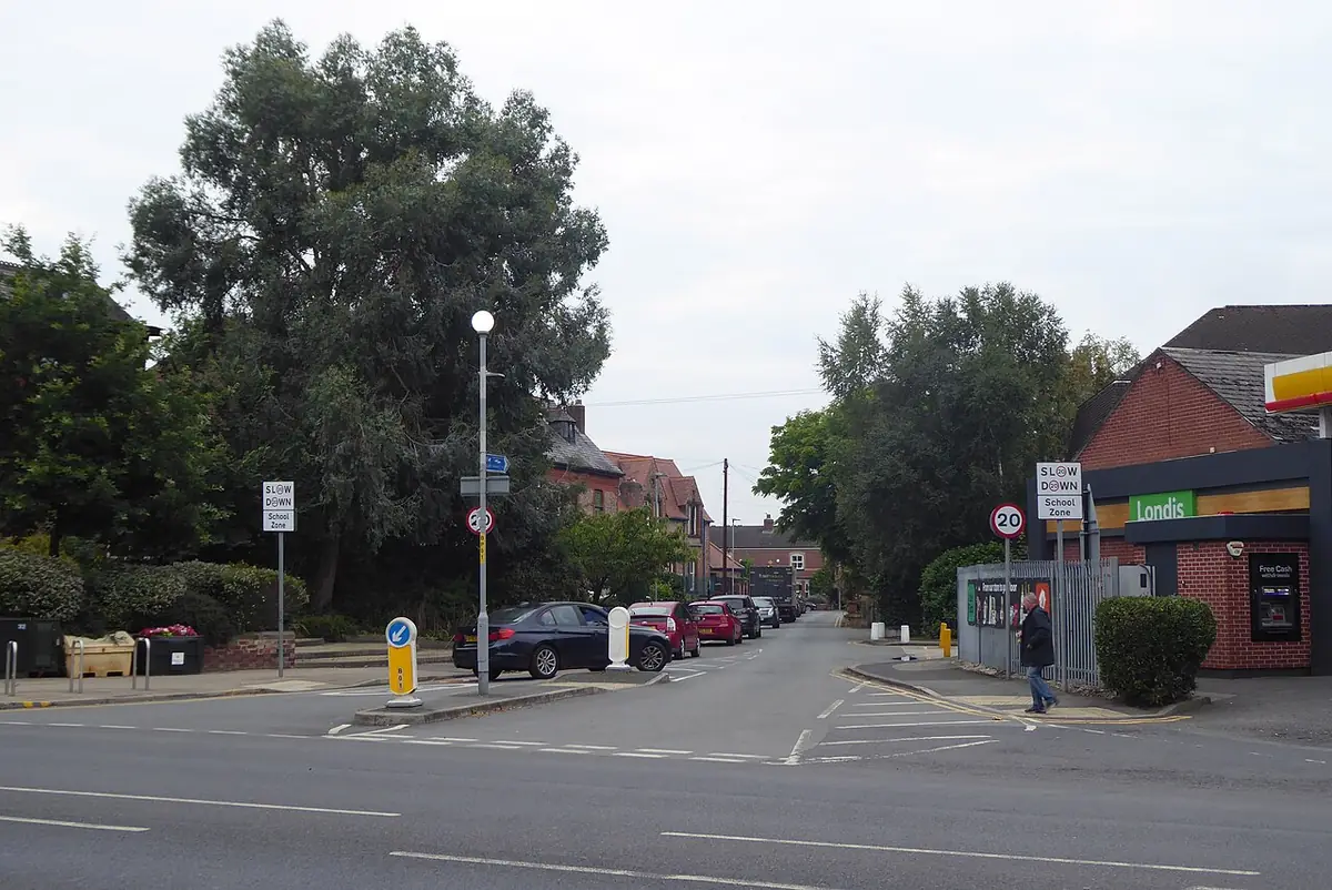 Un coin de rue à Manchester avec des arbres, des voitures garées et une supérette d’angle