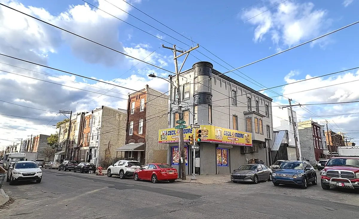 A Philadelphia street lined with rowhouses and corner shops under a partly cloudy sky