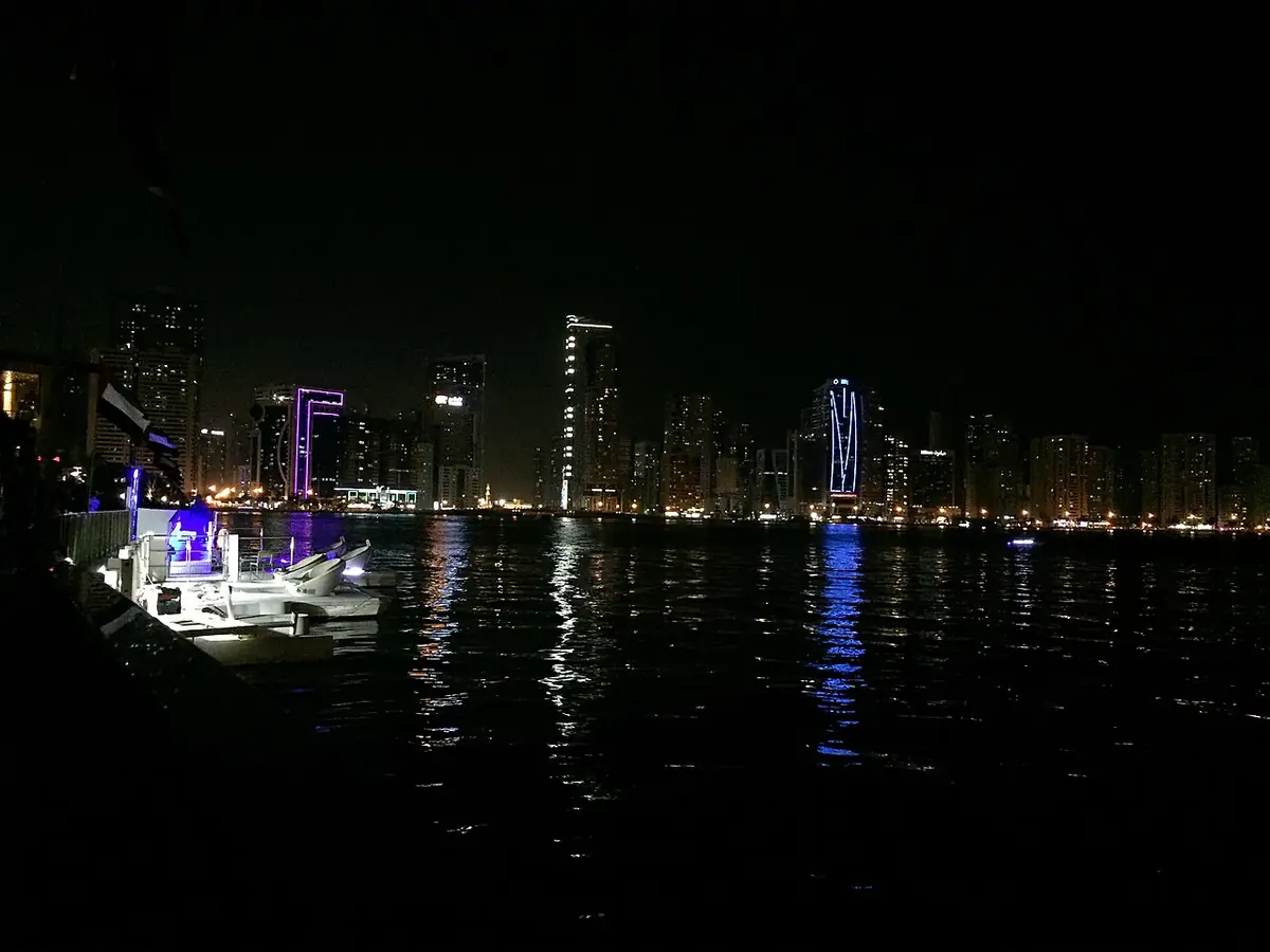 Des bateaux amarrés sur une eau sombre, la ligne d’horizon éclairée de Sharjah au loin